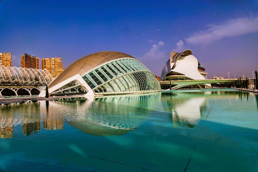 The futuristic architecture of the City of Arts and Sciences in Valencia, Spain, including the Hemisfèric and Palau de les Arts Reina Sofía, is reflected in a large pool of turquoise water under a clear blue sky.