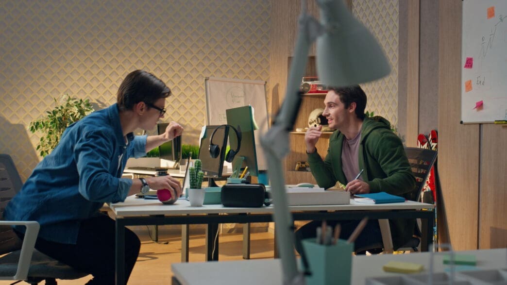 Two young men collaborating at a desk with computers in a modern office, one looking over at the other's screen.