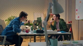 Two young men collaborating at a desk with computers in a modern office, one looking over at the other's screen.