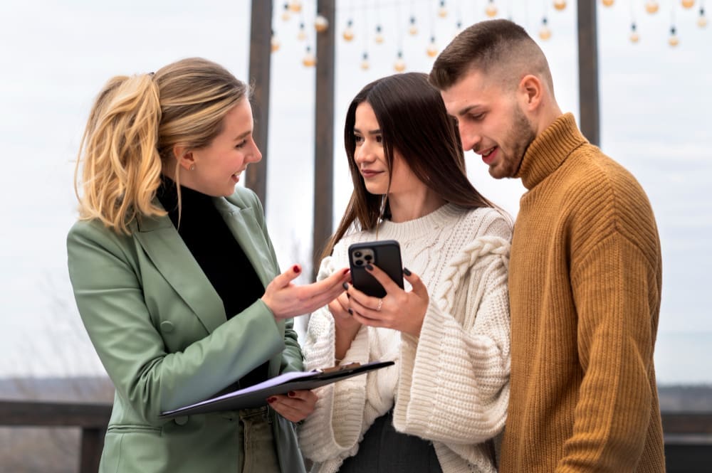 Three young adults, two women and one man, are engaged in a discussion while looking at a smartphone held by one of the women. The woman on the left is gesturing as she speaks.