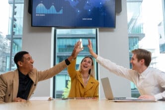 Three smiling diverse business professionals, two men and one woman, give each other high-fives across a table in a modern meeting room with a large screen displaying charts and a map.