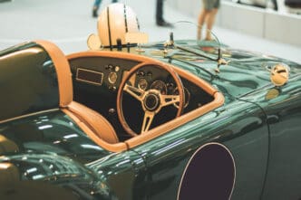 A close-up view of the cockpit of a vintage dark green convertible sports car with tan leather interior, a wooden steering wheel, and a white racing helmet on the seat.