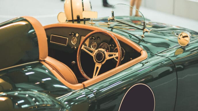 A close-up view of the cockpit of a vintage dark green convertible sports car with tan leather interior, a wooden steering wheel, and a white racing helmet on the seat.