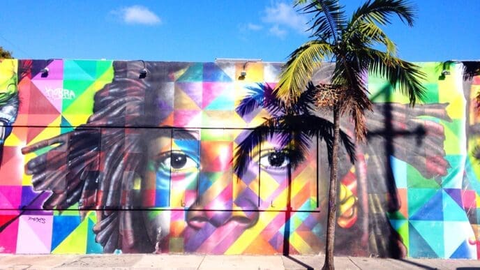 A vibrant street art mural on a building wall in Wynwood, Miami, depicting a large, colorful face with dreadlocks in a geometric pattern, partially obscured by the shadow of a palm tree, under a bright blue sky.