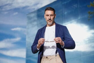 A confident, stylish man with a beard and mustache, wearing a blue blazer, white t-shirt, and suspenders over beige pants, holding eyeglasses, against a modern glass building and blue sky.