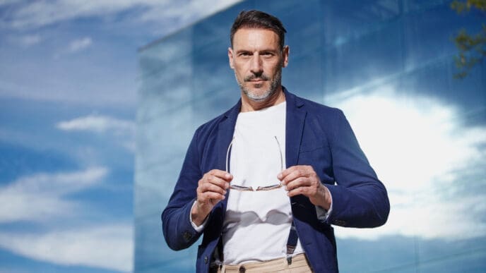 A confident, stylish man with a beard and mustache, wearing a blue blazer, white t-shirt, and suspenders over beige pants, holding eyeglasses, against a modern glass building and blue sky.