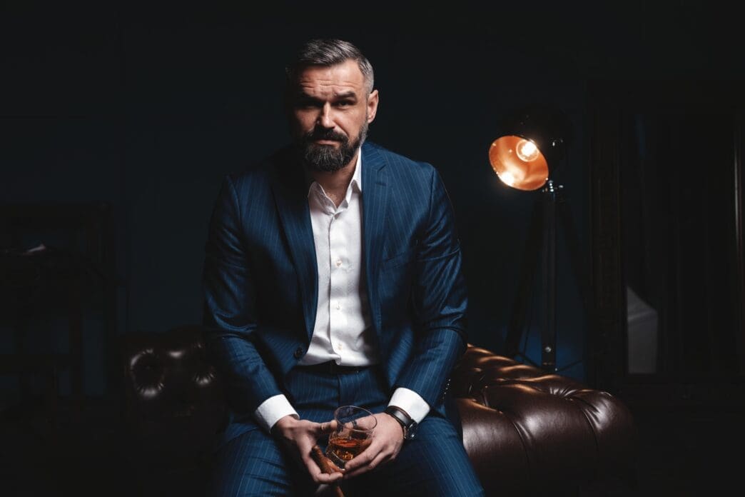 A serious, bearded man in a pinstripe suit, holding a glass of amber liquid, looking intently at the camera in a dimly lit, luxurious setting.