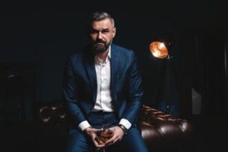 A serious, bearded man in a pinstripe suit, holding a glass of amber liquid, looking intently at the camera in a dimly lit, luxurious setting.