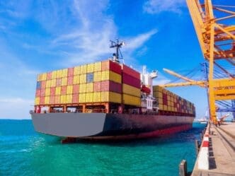 A large container ship, fully loaded with red and yellow containers, is docked at a port with orange gantry cranes visible on the right, under a bright blue sky.