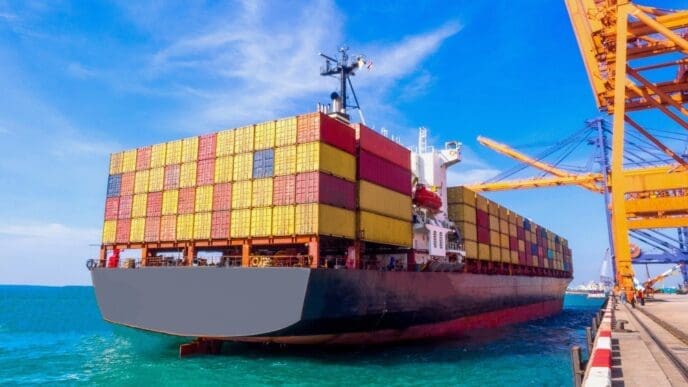 A large container ship, fully loaded with red and yellow containers, is docked at a port with orange gantry cranes visible on the right, under a bright blue sky.