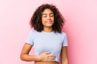 A young woman with curly hair, eyes closed and smiling serenely, holding her hand over her stomach, against a pink background.