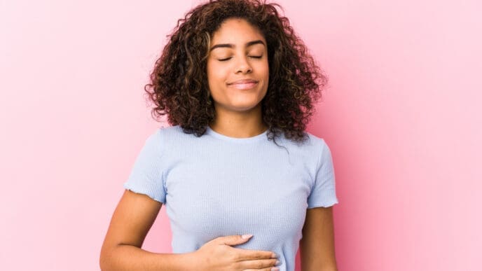 A young woman with curly hair, eyes closed and smiling serenely, holding her hand over her stomach, against a pink background.