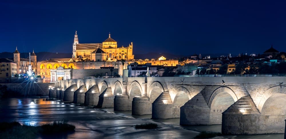 The illuminated Roman Bridge of Cordoba with its numerous arches spans a river at night, leading to the brightly lit Mezquita-Cathedral and the cityscape beyond, under a deep blue sky.