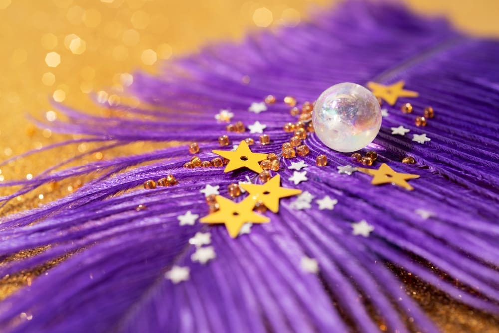 A close-up shot of a vibrant purple feather adorned with golden star confetti, tiny golden beads, and white glitter stars, with a clear iridescent sphere resting on it, all against a blurred golden glitter background.