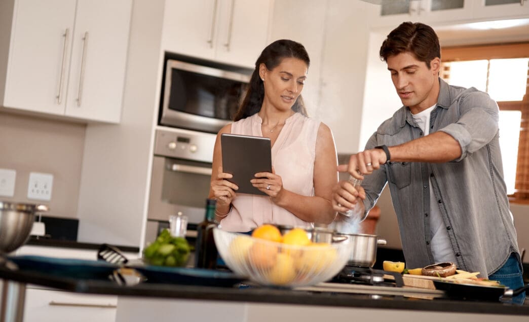 A cheerful couple cooking together in a modern kitchen, following a recipe on a digital tablet.