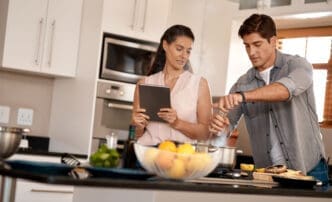 A cheerful couple cooking together in a modern kitchen, following a recipe on a digital tablet.
