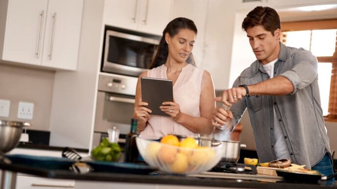 A cheerful couple cooking together in a modern kitchen, following a recipe on a digital tablet.