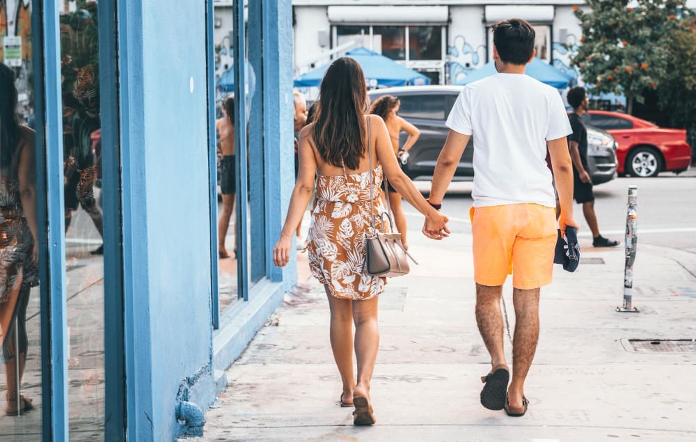 A couple walks hand-in-hand down a sidewalk in Wynwood, Miami. The woman wears a floral dress and carries a purse, while the man wears a white t-shirt and orange shorts. Reflective blue storefronts and street life are visible in the background.