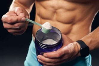 A muscular male torso is visible as hands scoop white powder, likely creatine, from a purple tub with a green scoop.
