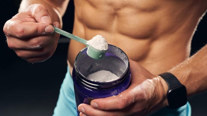 A muscular male torso is visible as hands scoop white powder, likely creatine, from a purple tub with a green scoop.