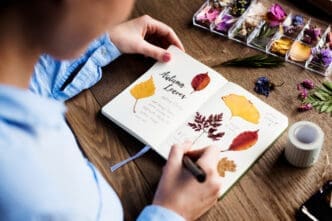 A person in a blue shirt writes in a nature journal filled with pressed autumn leaves and handwritten notes, with a clear organizer box of dried flowers and washi tape on a wooden table beside them.
