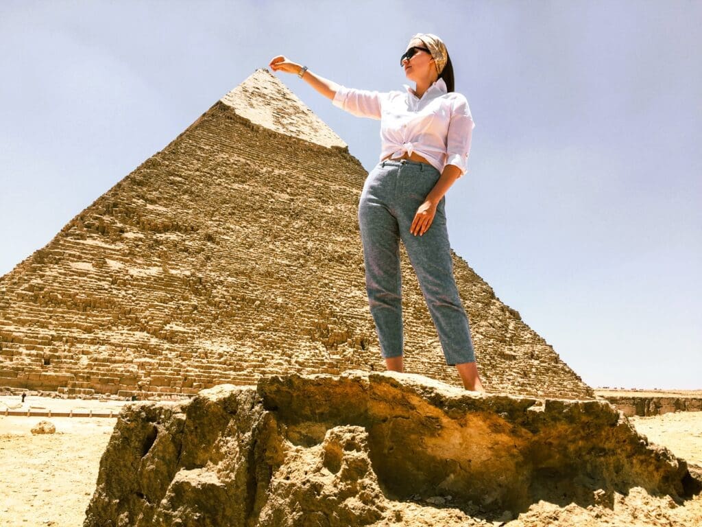 A young woman posing creatively with the Great Pyramid of Giza in the background, making it appear as if she is touching the top.