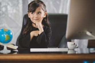 A young girl with dark hair and bangs, wearing a black shirt, sits at a large desk with a computer monitor, globe, and mug, looking thoughtfully to the side with her hand on her chin.