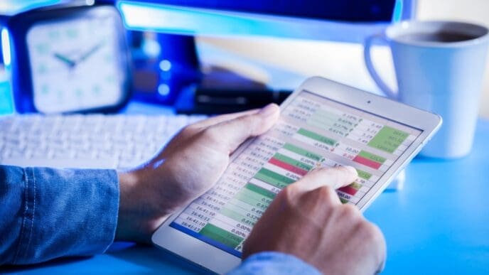 A close-up of a person's hands holding a digital tablet that displays a complex financial spreadsheet or budgeting data, with a finger touching the screen.