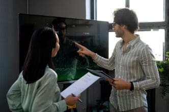 Two professionals, a man and a woman, are discussing data displayed on a large screen; the man points to the screen while holding documents, and the woman holds papers, looking at him.