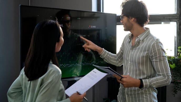 Two professionals, a man and a woman, are discussing data displayed on a large screen; the man points to the screen while holding documents, and the woman holds papers, looking at him.