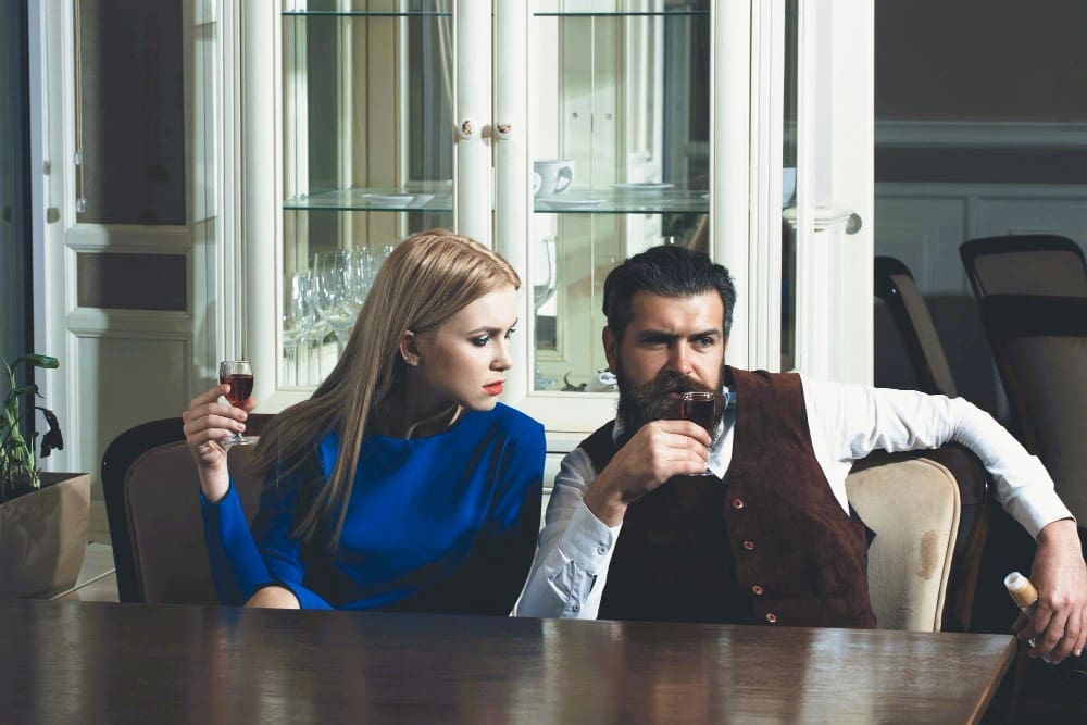 A serious-looking man with a beard and a woman with blonde hair sit at a table, both holding glasses of red wine, with the man looking thoughtful and the woman looking towards him, suggesting a deep conversation in a formal setting.