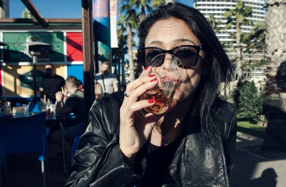 A stylish woman in a black leather jacket and sunglasses sips a drink from a glass at an outdoor cafe. The background shows palm trees and modern buildings, suggesting a sunny day in Miami's Wynwood neighborhood.