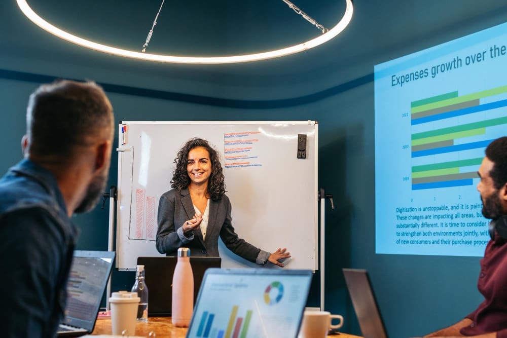 A smiling female presenter in a grey suit stands in front of a whiteboard with charts, gesturing to her audience, who are visible from behind with laptops, in a modern meeting room with a large projected screen showing financial data.