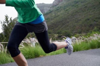 A close-up of a runner's legs in activewear and running shoes, captured mid-stride on an outdoor path with green hills in the background.