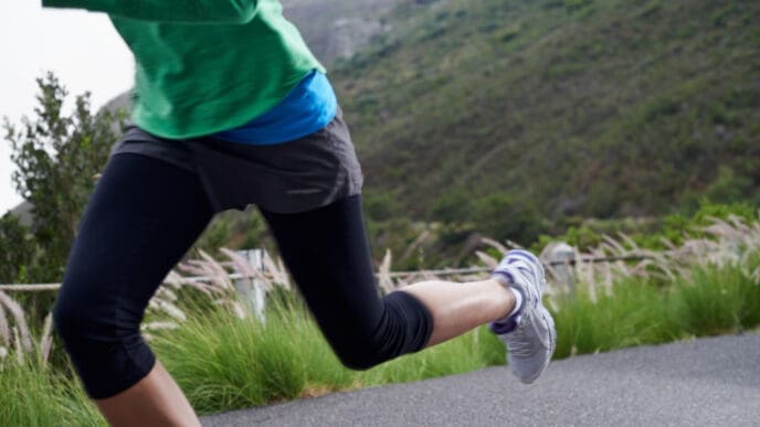 A close-up of a runner's legs in activewear and running shoes, captured mid-stride on an outdoor path with green hills in the background.