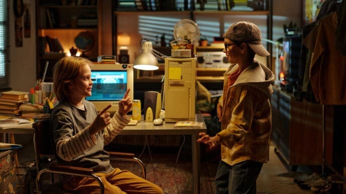 Two young boys in a cluttered room with an old computer, engaged in conversation, reminiscent of early tech startup environments.