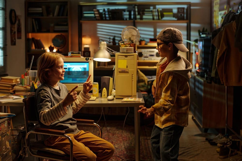 Two young boys in a cluttered room with an old computer, engaged in conversation, reminiscent of early tech startup environments.