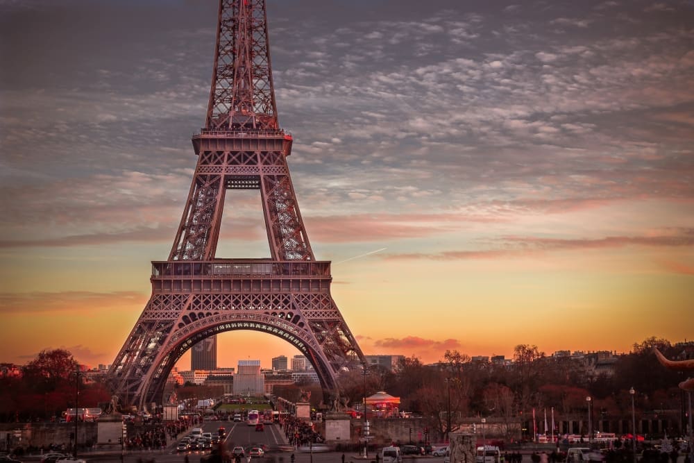 A low-angle view of the Eiffel Tower in Paris, France, against a vibrant sunset sky, showing its intricate iron lattice structure and the surrounding cityscape.