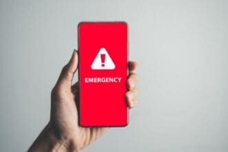 A hand holds a smartphone displaying a bright red screen with a large white warning triangle and the word "EMERGENCY," symbolizing a crisis or urgent situation.