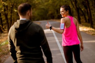 A woman in a pink tank top and a man in a dark hoodie walk or jog on a paved road through a wooded area. The woman looks over her shoulder, smiling and giving a thumbs-up gesture, wearing a phone armband.