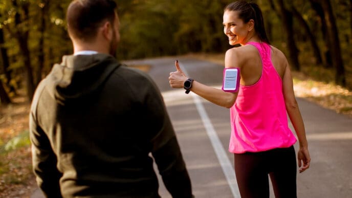 A woman in a pink tank top and a man in a dark hoodie walk or jog on a paved road through a wooded area. The woman looks over her shoulder, smiling and giving a thumbs-up gesture, wearing a phone armband.