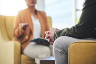 A close-up on a person's clasped hands during a therapy session, with a therapist gesturing in the blurred background.