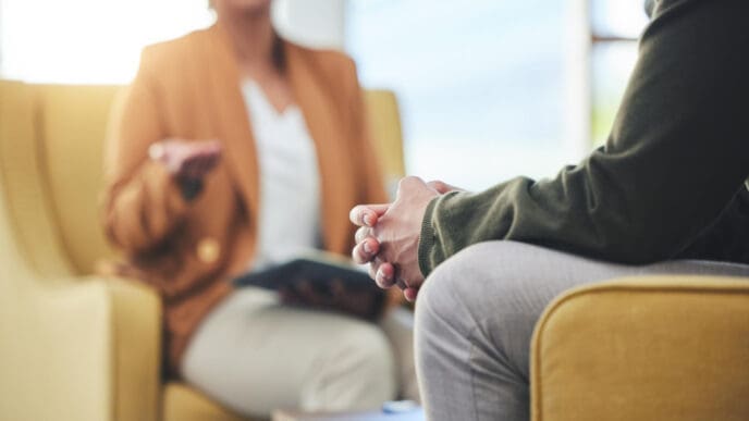 A close-up on a person's clasped hands during a therapy session, with a therapist gesturing in the blurred background.