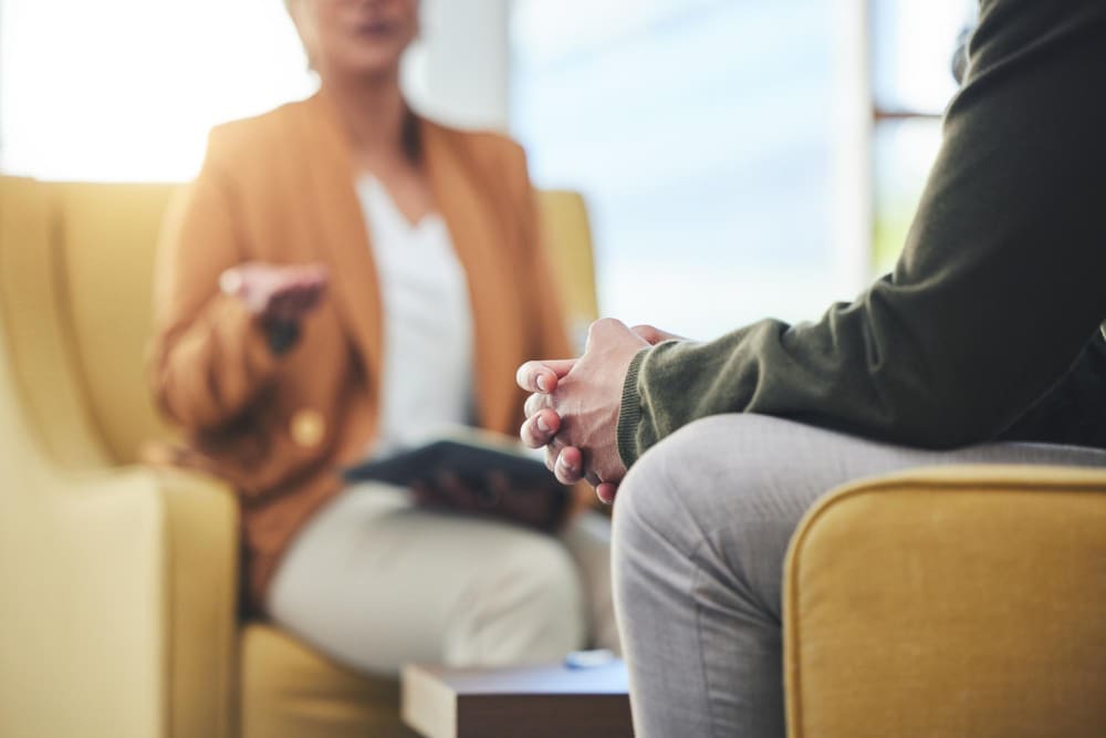 A close-up on a person's clasped hands during a therapy session, with a therapist gesturing in the blurred background.