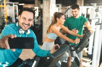 A smiling man taking a selfie with headphones while on an exercise bike, with a woman and trainer in the background at a gym.