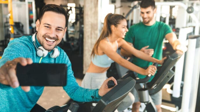 A smiling man taking a selfie with headphones while on an exercise bike, with a woman and trainer in the background at a gym.