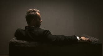 A man in a suit, seen from behind, sitting on a couch in a dimly lit room, with his face in profile.