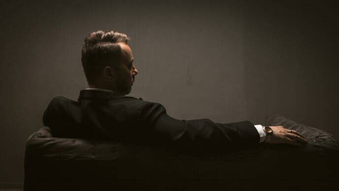 A man in a suit, seen from behind, sitting on a couch in a dimly lit room, with his face in profile.