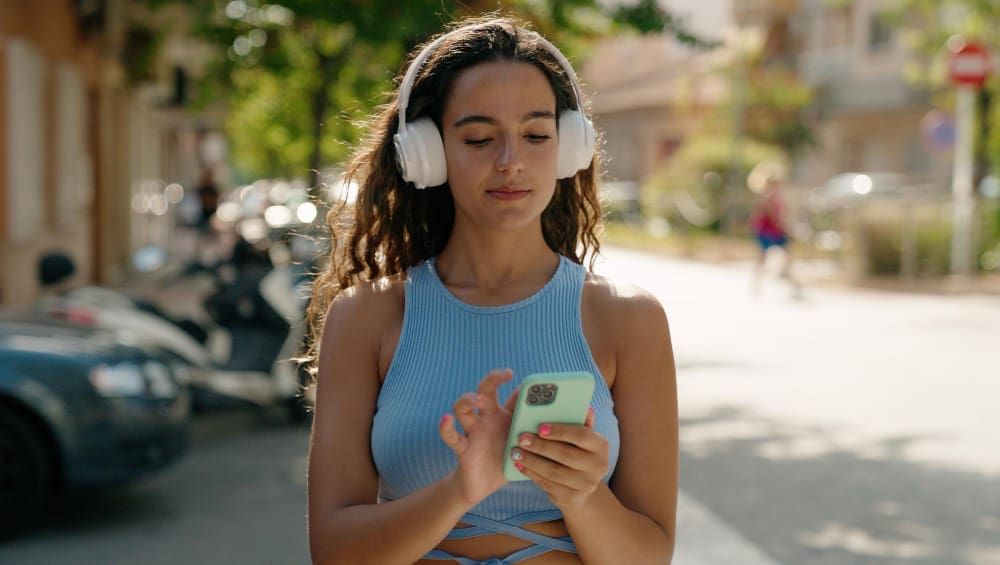 A young woman with long curly hair, wearing white headphones and a light blue top, walks outdoors while looking at and operating her light green smartphone.