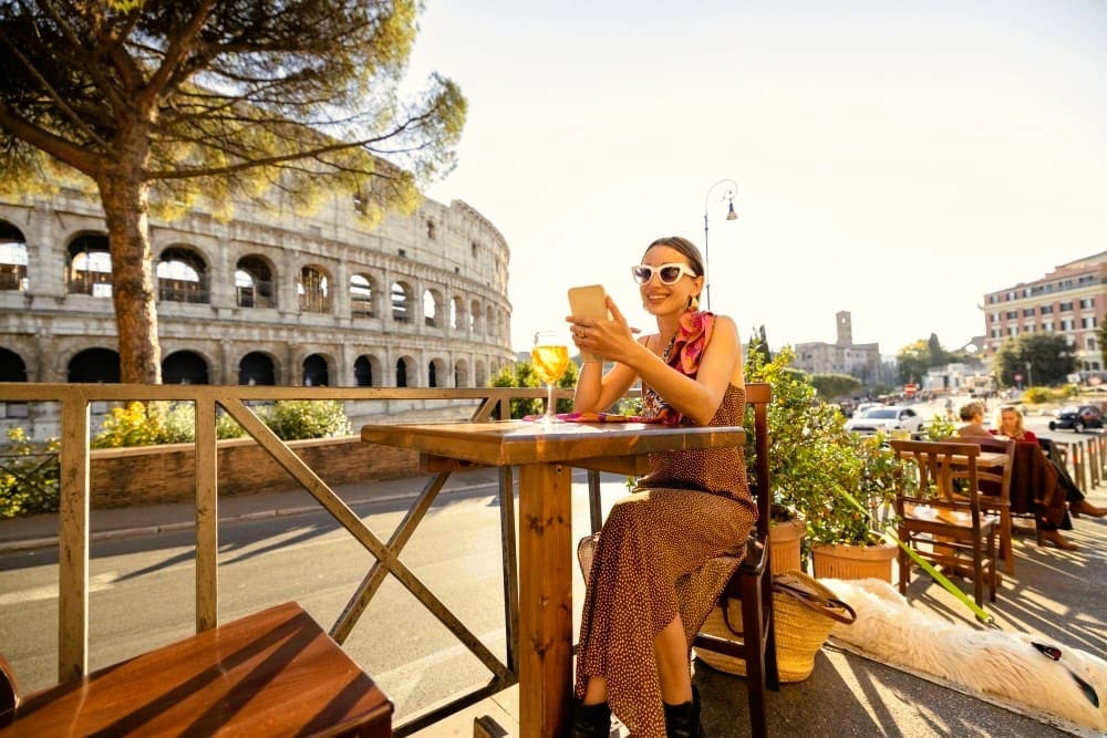 A young woman, smiling and wearing sunglasses, sits at an outdoor cafe table looking at her smartphone. The ancient Roman Colosseum is visible in the background under a sunny sky.